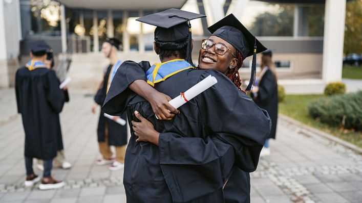Male and female student in graduation gowns embracing each other on the university campus outdoors.